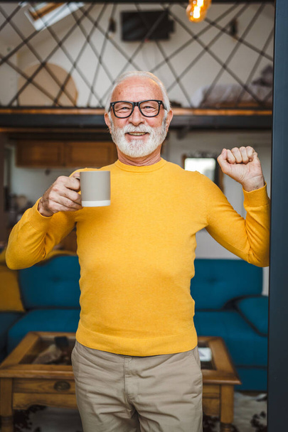 One man senior caucasian male with beard and eyeglasses stand on balcony at door in day happy smile hold cup of coffee copy space morning daily routine concept - Fotó, kép