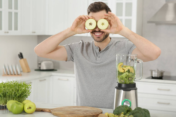 Man covering eyes with halves of apples in kitchen. Making smoothie - 写真・画像