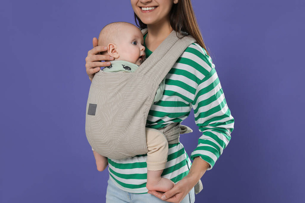 Mother holding her child in sling (baby carrier) on purple background, closeup - Foto, imagen