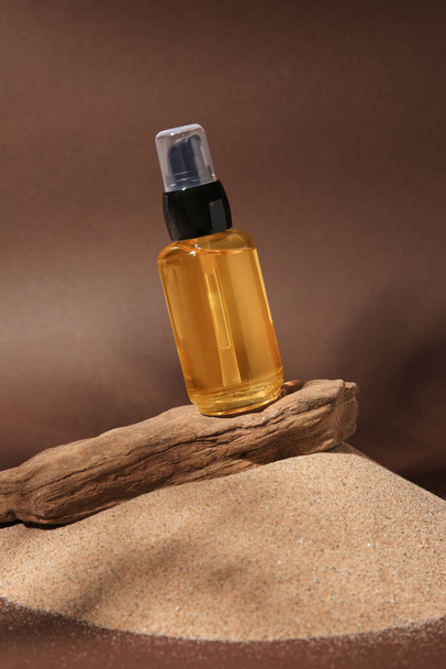Bottle of serum and tree bark on sand against brown background - Фото, изображение