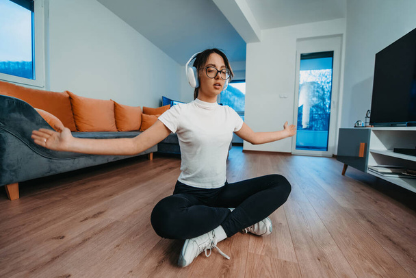 Genç bir kadın yoga yapıyor ve kafasında kulaklıklarla modern bir dairede dinleniyor.. - Fotoğraf, Görsel