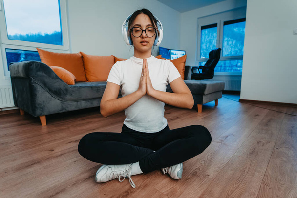 Eine junge Frau praktiziert Yoga und entspannt sich mit Kopfhörern auf dem Kopf in einer modernen Wohnung. - Foto, Bild
