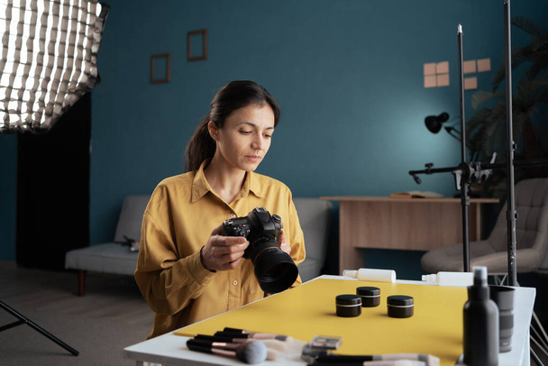 Fotografa donna mettendo prodotti cosmetici in tavola facendo composizione, facendo foto del prodotto di trucco, tenendo la fotocamera in studio di casa. blogger moderno. Creatore di contenuti. Copia spazio - Foto, immagini