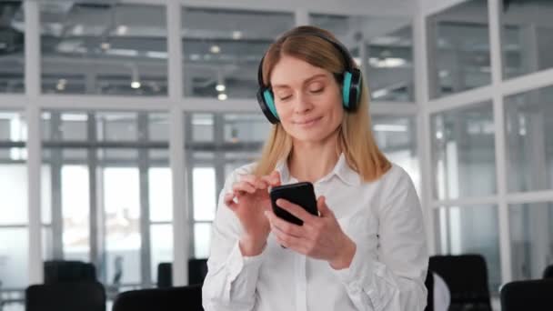 Funny Businesswoman Dancing In Wireless Headphones With Mobile phone in Office. Žena ilustrující, jak může být práce užitečná, inspirující tvořivost, spolupráce a rovnováha mezi pracovním a soukromým životem - Záběry, video