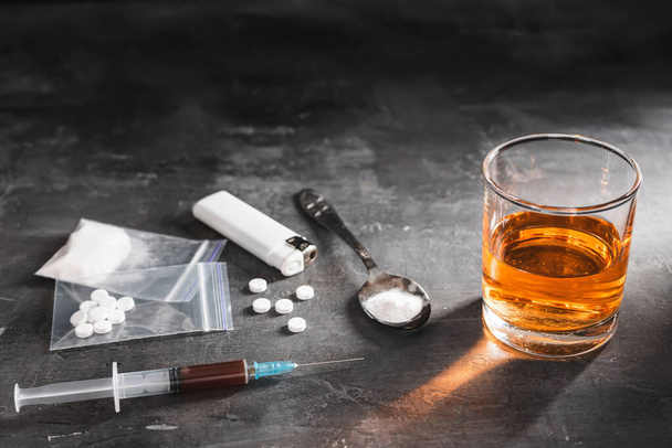 Alcohol drink in a glass, syringe with a dose of drugs, white pills in a transparent bag and narcotics powder in a spoon on dark background. Concept of addiction and bad habits. - Фото, зображення