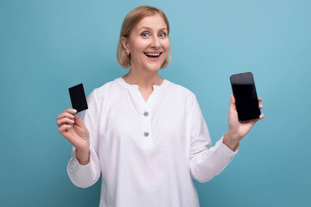 soort aangename blonde volwassen vrouw met kaart en smartphone voor winkelen met mok op studio achtergrond. - Foto, afbeelding