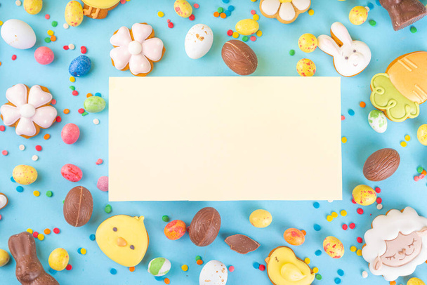 Easter sweets and candies background. High-colored blue flat lay with various Easter chocolate eggs, treats, symbols of easter sheep, bunny, carrot, flowers, cookies, top view copy space, frame - Zdjęcie, obraz