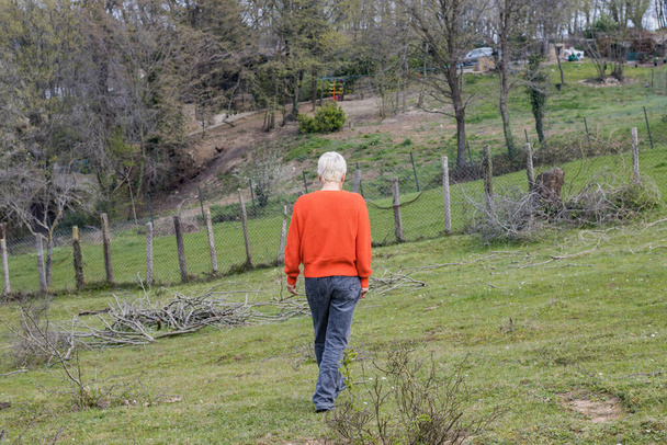blonde woman in orange walking away into grassland with a farm on it - Φωτογραφία, εικόνα