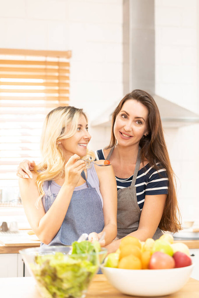 Glückliches gleichgeschlechtliches Paar füttert einander in der Küche. Fröhliche LGBTQ-Lesben füttern sich gegenseitig mit Salat. Ein schönes Paar lesbischer Damen hat einen romantischen Moment in der Küche. - Foto, Bild
