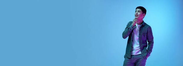 Schöner junger asiatischer Mann posiert in lässiger Kleidung mit nachdenklichem, träumerischem Gesicht vor blauem Hintergrund im Neonlicht. Konzept menschlicher Emotionen, Jugend, Mode, Lebensstil. Banner. Kopierraum für Werbung - Foto, Bild