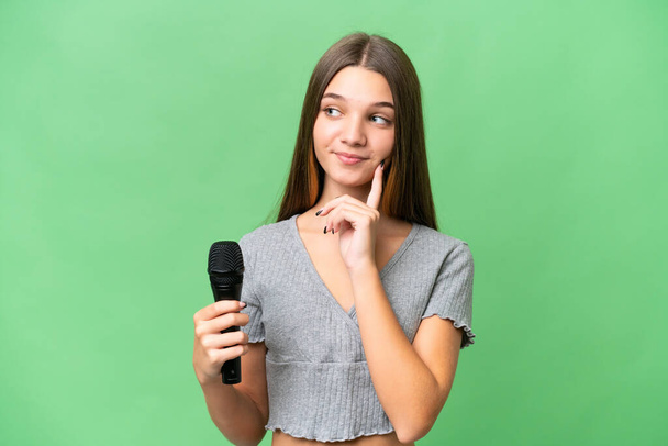 Ragazza cantante adolescente raccogliendo un microfono su sfondo isolato pensando un'idea mentre guardando in su - Foto, immagini
