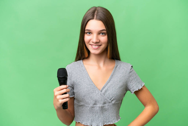Подросток певица поднимает микрофон на изолированном фоне позирует с руками на бедре и улыбается - Фото, изображение