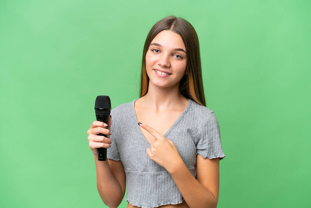 Genç şarkıcı kız izole edilmiş bir arka planda mikrofonu kaldırıyor ve bir ürünü sunmak için kenarı işaret ediyor. - Fotoğraf, Görsel