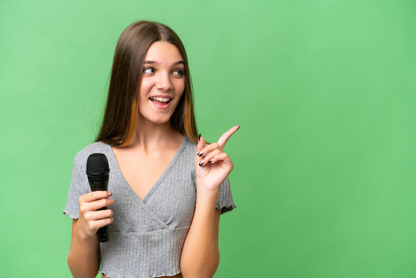Tiener zangeres meisje het oppakken van een microfoon over geïsoleerde achtergrond van plan om de oplossing te realiseren, terwijl het opheffen van een vinger omhoog - Foto, afbeelding