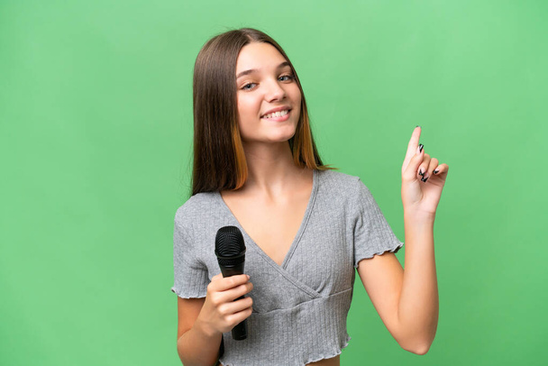 Adolescente cantora menina pegar um microfone sobre fundo isolado mostrando e levantando um dedo em sinal do melhor - Foto, Imagem