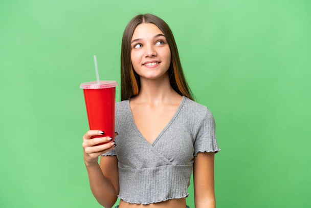 Adolescente caucásico chica sosteniendo un refresco sobre aislado fondo mirando hacia arriba mientras sonríe - Foto, Imagen