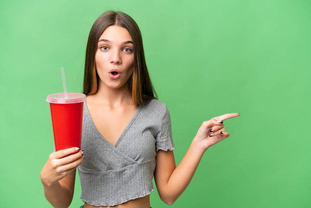 Teenager caucasian girl holding a soda over isolated background surprised and pointing side - Фото, зображення
