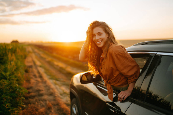 Young happy woman leaning out of the car window enjoying the sunset. The concept of active lifestyle, travel, tourism, nature. - 写真・画像