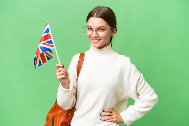 Подростковая студентка кавказская девушка с флагом Великобритании на изолированном фоне позирует с руками на бедре и улыбается - Фото, изображение