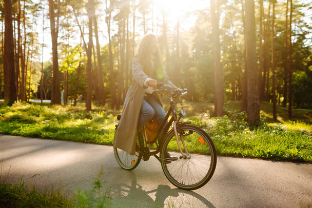 Smiling woman with curly hair in a coat rides a bicycle in a sunny park. Outdoor portrait. Beautiful woman enjoys nature. Lifestyle. Relax, nature concept.  - Foto, Bild