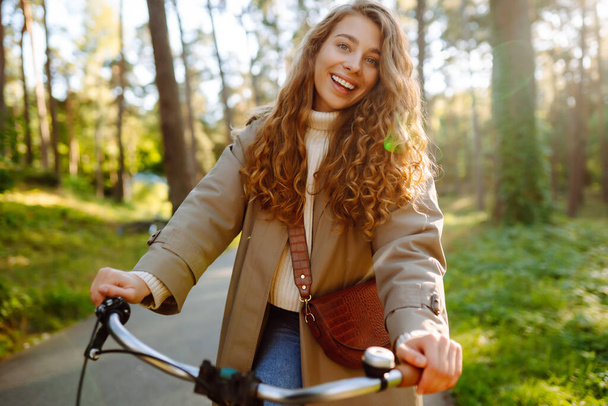 Smiling woman with curly hair in a coat rides a bicycle in a sunny park. Outdoor portrait. Beautiful woman enjoys nature. Lifestyle. Relax, nature concept.  - Fotó, kép