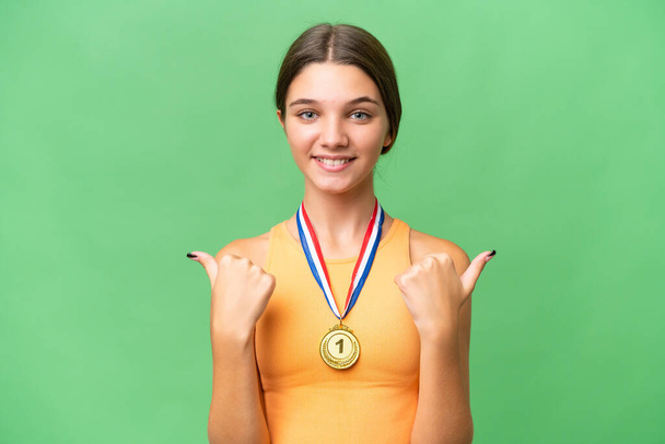 Adolescente caucasienne avec des médailles sur fond isolé avec les pouces vers le haut geste et sourire - Photo, image