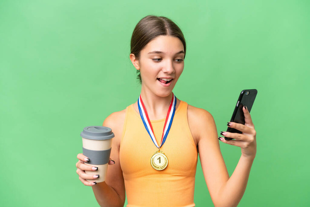 Teenager caucasian girl with medals over isolated background holding coffee to take away and a mobile - Foto, afbeelding