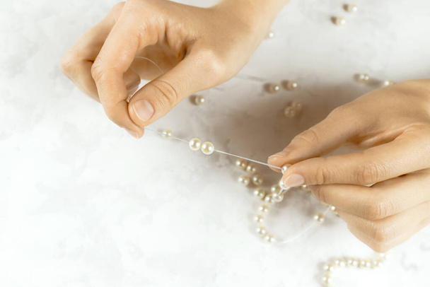 Close-up of woman hands stringing white pearls on a precious necklace on white background. Beads jewellery making process. Hobby handmade concept. Top down view. Selective focus. Blurred background. - Фото, зображення