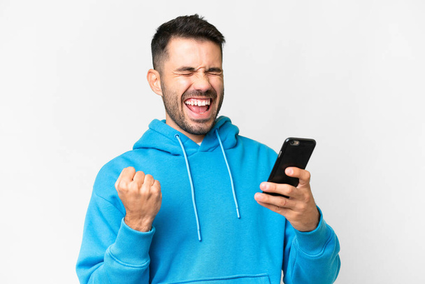 Young handsome caucasian man over isolated white background with phone in victory position - Zdjęcie, obraz