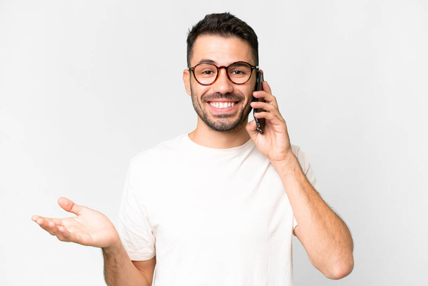 Молодой красивый кавказский мужчина на изолированном белом фоне ведет разговор с кем-то по мобильному телефону - Фото, изображение