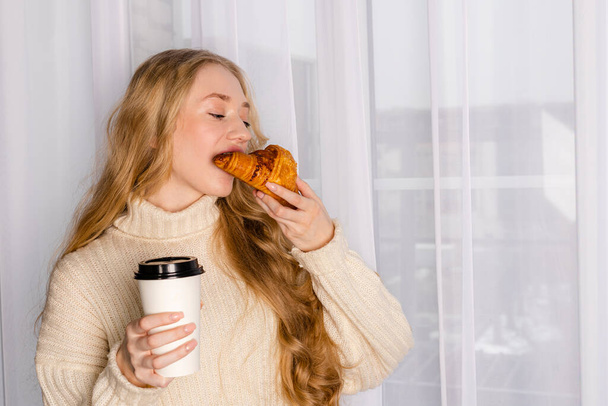 Young tender beautiful long haired girl in a sweater standing against white curtains with a blank white paper coffee cup and a tasty croissant. - Foto, immagini