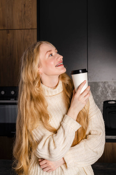 Jeune fille tendre et belle aux cheveux longs dans un pull et un jean assis sur une table de cuisine moderne avec une maquette de tasse à café en papier blanc. - Photo, image