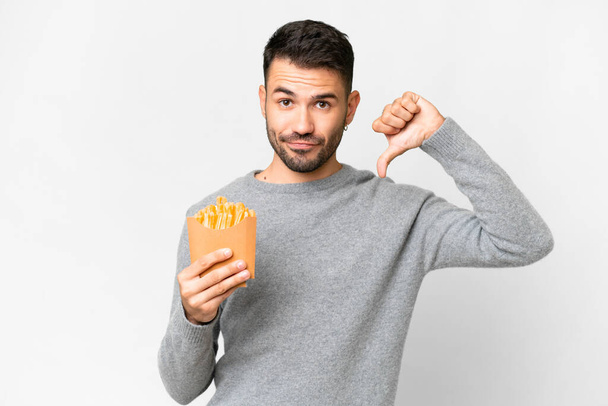 Jonge blanke man met gebakken chips over geïsoleerde witte achtergrond met duim naar beneden met negatieve expressie - Foto, afbeelding
