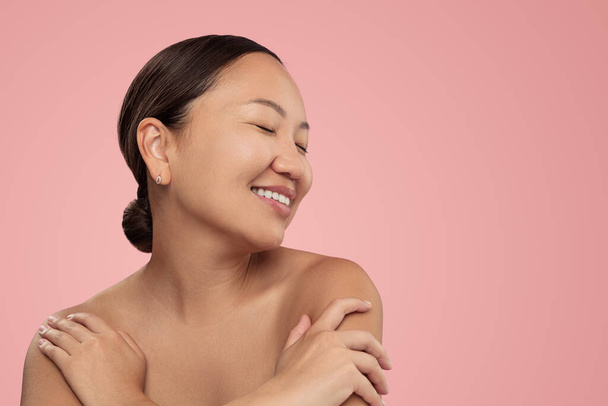 Smiling ethnic female with bare shoulders closed eyes and dark hair standing against pink background embracing herself - Photo, Image