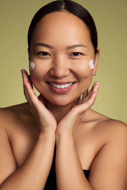 Asian female model with bare shoulders applying moisturizing cream on face while looking at camera against green background - Foto, Bild