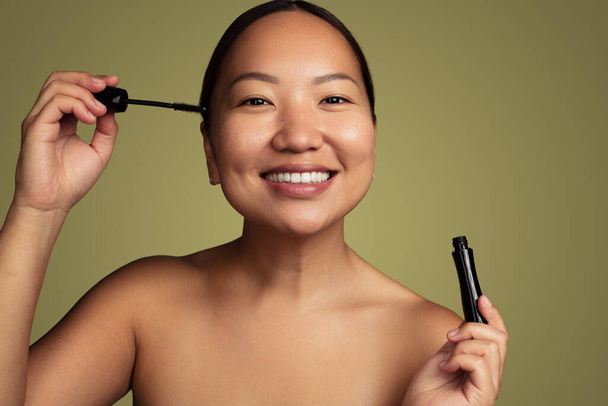 Cheerful young ethnic female with bare shoulders smiling and looking at camera while applying black mascara on eyelashes against green background - Foto, imagen