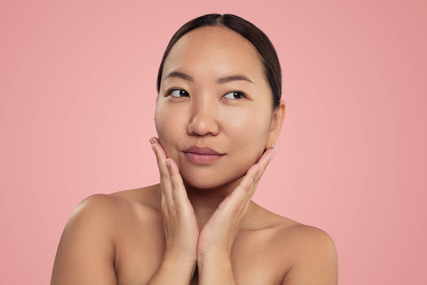 Young ethnic female with bare shoulders standing in studio after skincare routine looking away against pink background - Fotó, kép
