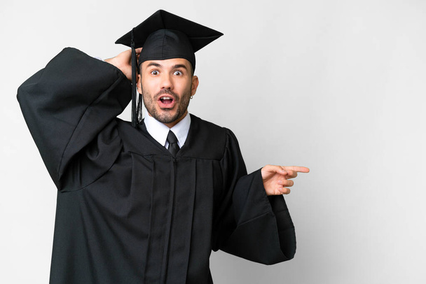Молодой выпускник университета на изолированном белом фоне удивлен и указывает пальцем в сторону - Фото, изображение