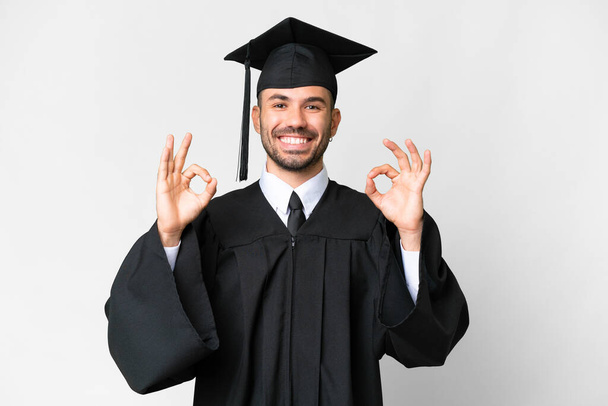 Молодой выпускник университета на изолированном белом фоне показывает нормальный знак с пальцами - Фото, изображение