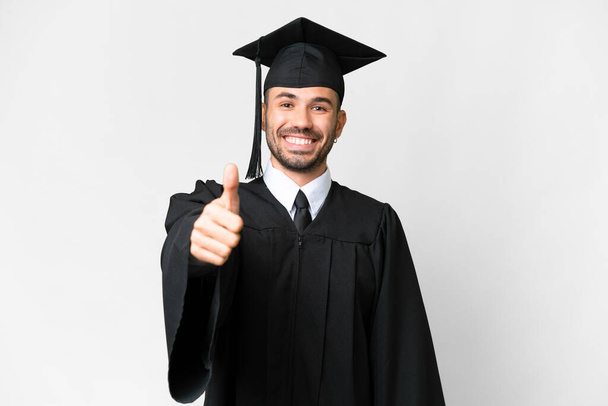 Fiatal egyetemi diplomás férfi elszigetelt fehér háttér hüvelykujjal felfelé, mert valami jó történt - Fotó, kép