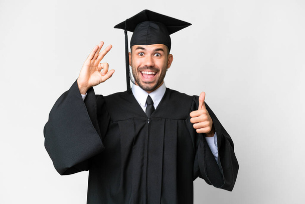 jonge universiteit afgestudeerde man over geïsoleerde witte achtergrond tonen ok teken en duim omhoog gebaar - Foto, afbeelding