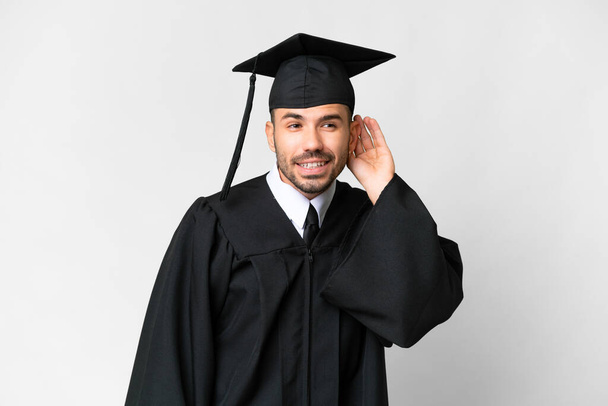 Молодой выпускник университета на изолированном белом фоне слушает что-то, прикладывая руку к уху - Фото, изображение