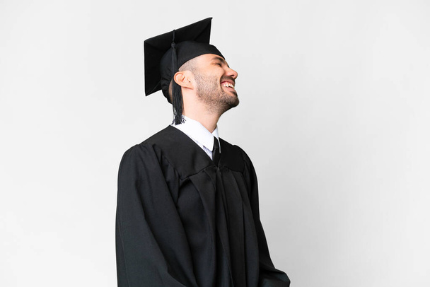 Jonge afgestudeerde man over geïsoleerde witte achtergrond lachen in laterale positie - Foto, afbeelding