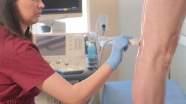 Egy ortopéd orvos ultrahangos vizsgálatot végez egy beteg térdén. Egy idős nőt ultrahang vizsgálatnak vetnek alá egy klinikán. Ultrahangos orvosi kutatás. 4k videó - Felvétel, videó