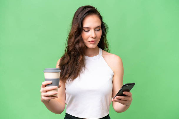 Young caucasian woman isolated over isolated background holding coffee to take away and a mobile - Фото, зображення