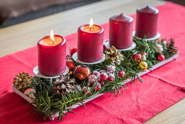 Burning red advent candles with decoration on a wooden table in living room on red fabric cloth at Christmas time, Germany - Photo, Image