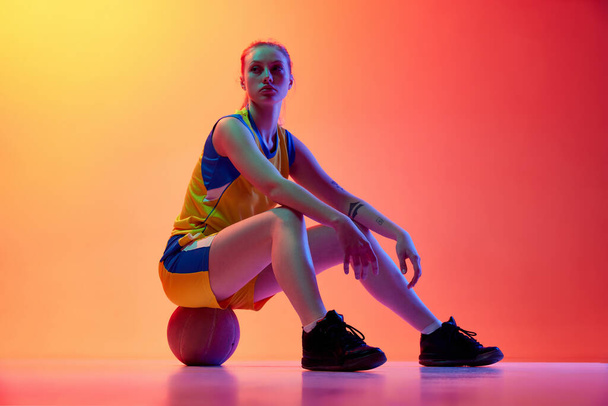 After game. Young girl, female basketball player in uniform, posing, sitting on ball against white studio background. Concept of professional sport, hobby, healthy lifestyle, action and motion - Φωτογραφία, εικόνα
