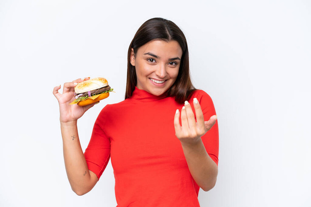 Mujer caucásica joven sosteniendo una hamburguesa aislada sobre fondo blanco invitando a venir con la mano. Feliz de que hayas venido. - Foto, Imagen