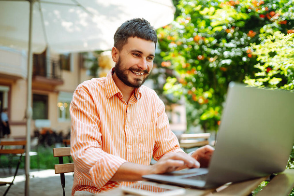 Χαμογελώντας νεαρός άνδρας που εργάζεται σε ένα καφέ στο δρόμο με ένα φορητό υπολογιστή και τηλέφωνο. Ανεξάρτητη επιχειρηματική ιδέα. Επιχειρήσεις, blogging, freelancing, εκπαιδευτική έννοια. Σύγχρονος τρόπος ζωής. - Φωτογραφία, εικόνα