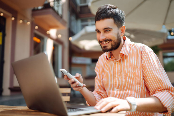 Een glimlachende jongeman in een café op straat met een laptop en telefoon. Freelance business concept. Zakelijk, bloggen, freelancen, onderwijs concept. Moderne levensstijl. - Foto, afbeelding